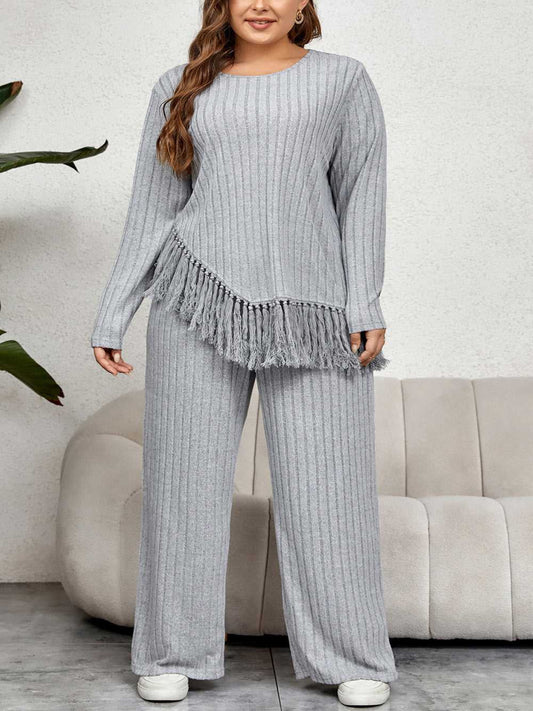 Woman wearing a gray ribbed sweater and pants with fringe detail, standing in a room with a couch and plant.